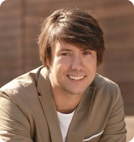Young man with medium-length brown hair smiling, wearing a beige blazer over a white shirt, with a blurred brown background.