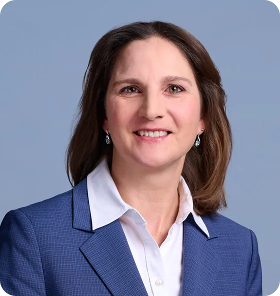 Smiling woman with shoulder-length brown hair wearing a blue blazer, white collared shirt, and teardrop earrings in front of a light blue background.