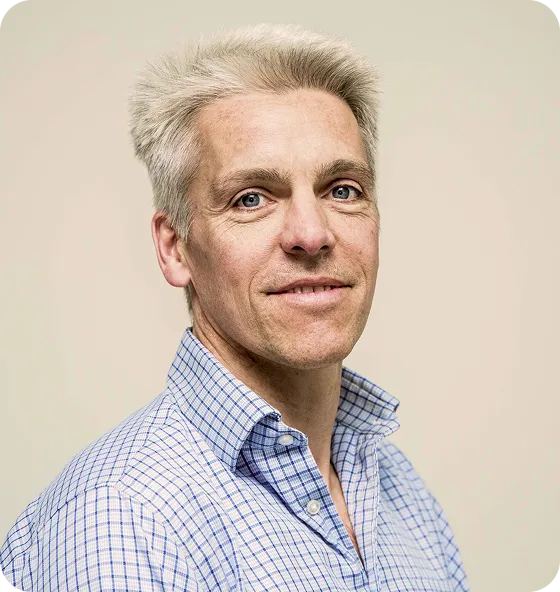 Portrait of a middle-aged man with short gray hair and blue eyes wearing a blue and white checkered button-up shirt against a plain beige background.