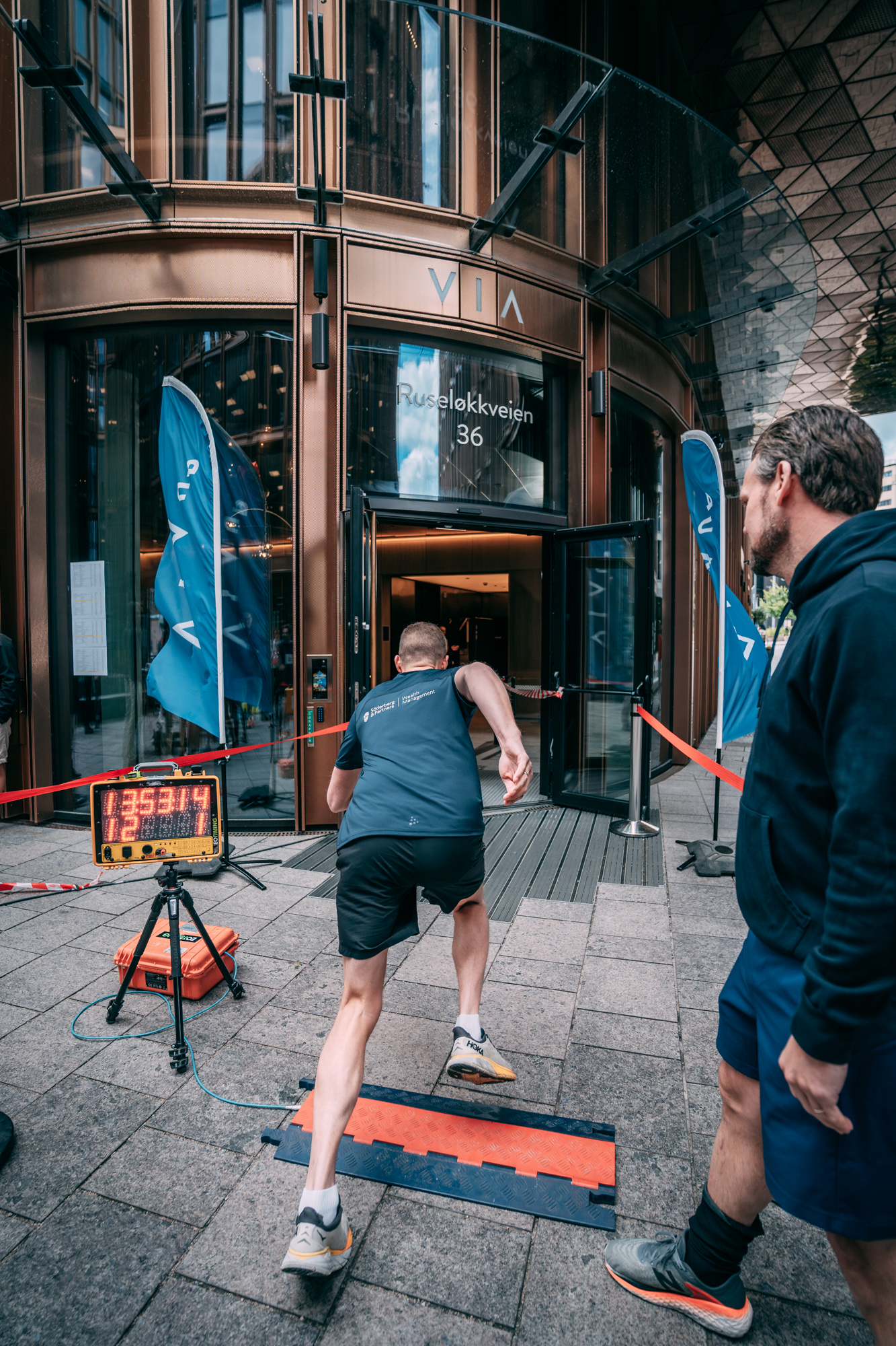Rear view of a man sprinting on a city sidewalk timer running while a man watches near building entrance.