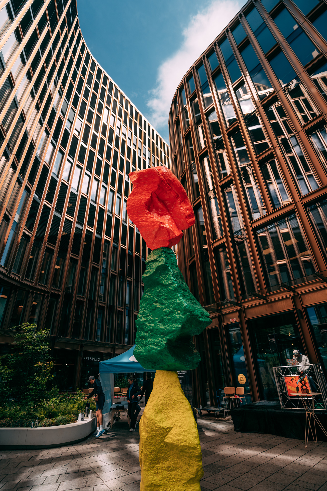 Colorful three-part rock sculpture with yellow, green, and red stones in a courtyard with modern glass buildings.