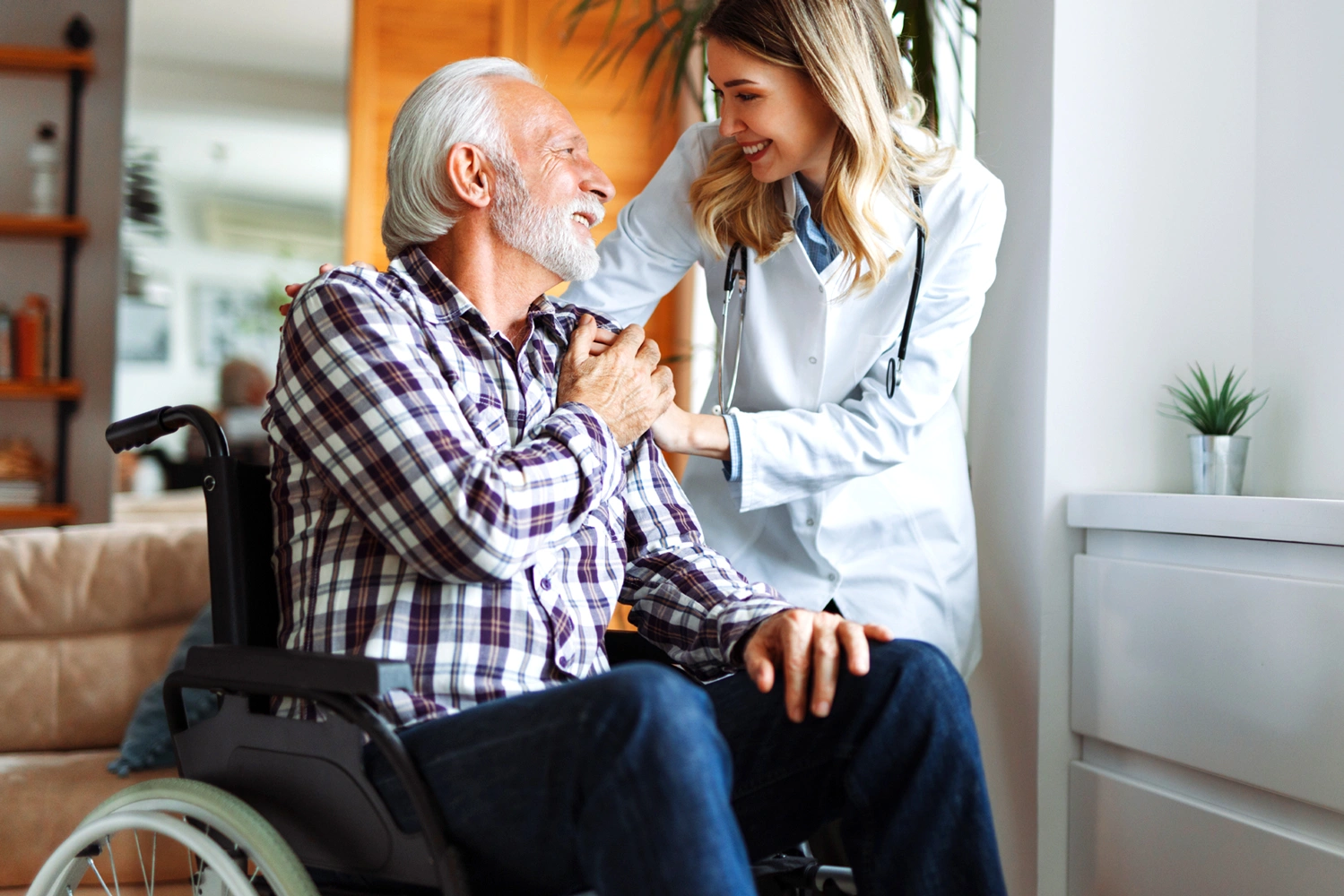 Une médecin souriante soutient un homme âgé assis en fauteuil roulant à la maison.
