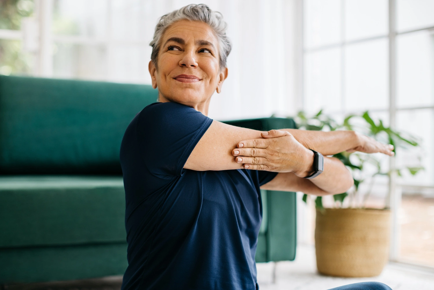 Une femme souriante en tenue de sport fait des étirements des bras dans un salon lumineux.