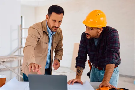 Deux hommes discutant d'un projet devant un ordinateur portable sur un chantier de construction.