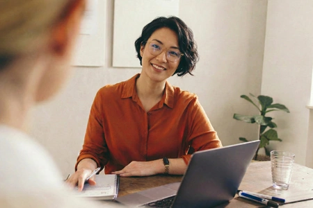 Femme souriante en chemise orange avec ordinateur portable et bloc-notes, parlant à une autre personne.
