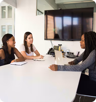Trois femmes discutent autour d'une table blanche dans une salle de réunion moderne avec un grand écran.