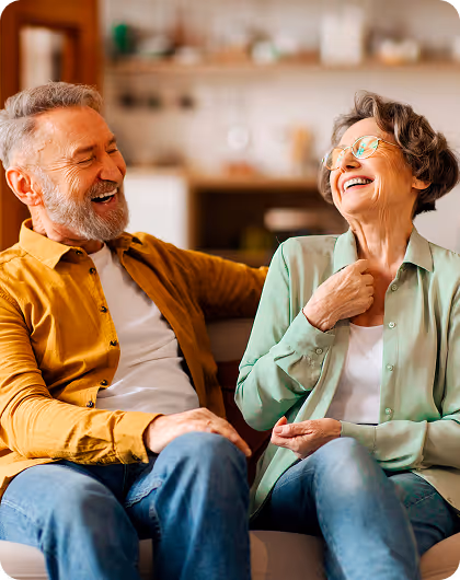 Un homme et une femme âgés souriant et riant ensemble assis dans un salon confortable.