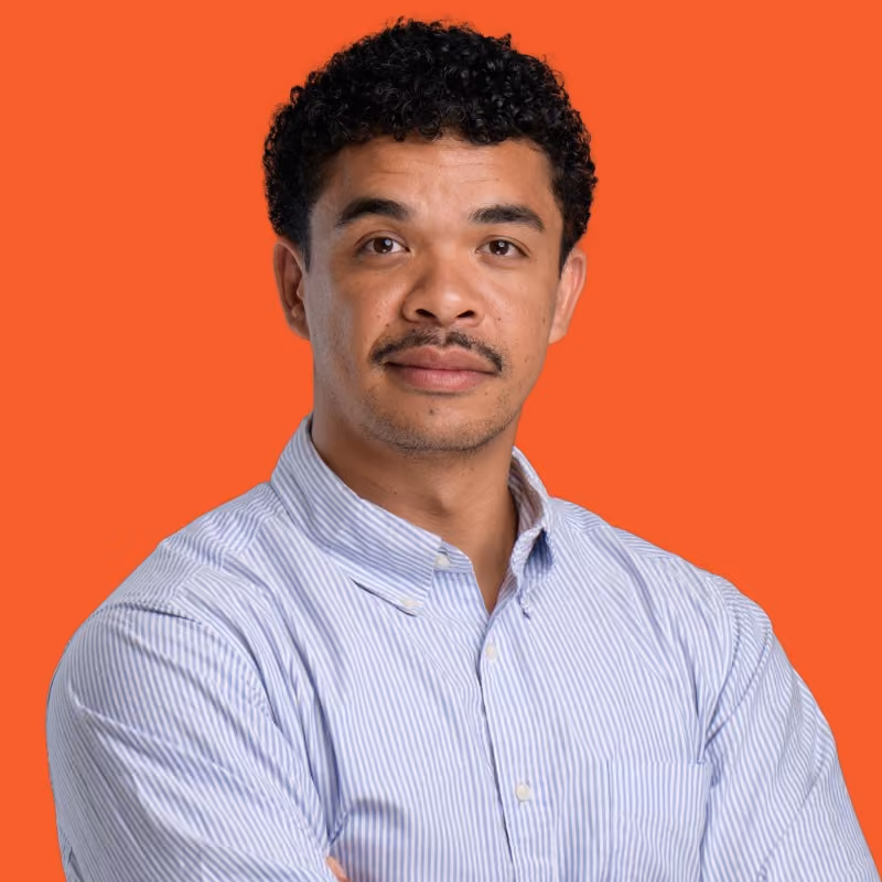 Homme souriant aux cheveux bouclés et moustache, portant un t-shirt noir sur fond orange.