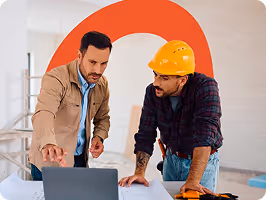 Two men discussing work while looking at a laptop, one wearing a yellow construction helmet.