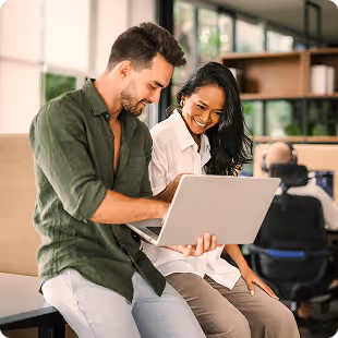 Deux collègues souriants regardent un ordinateur portable dans un bureau moderne.
