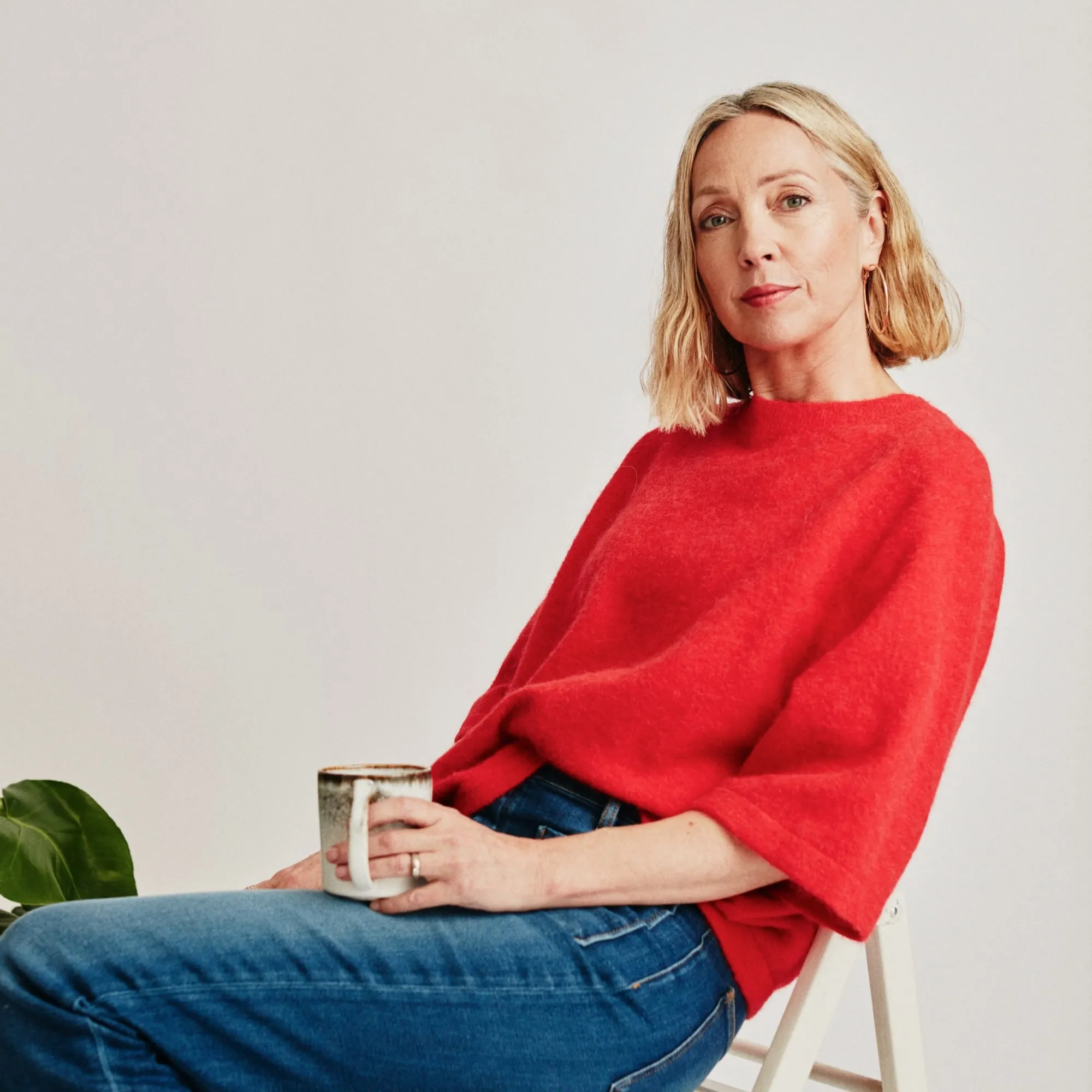 Woman with blonde hair in red sweater and jeans sitting on chair holding a mug.