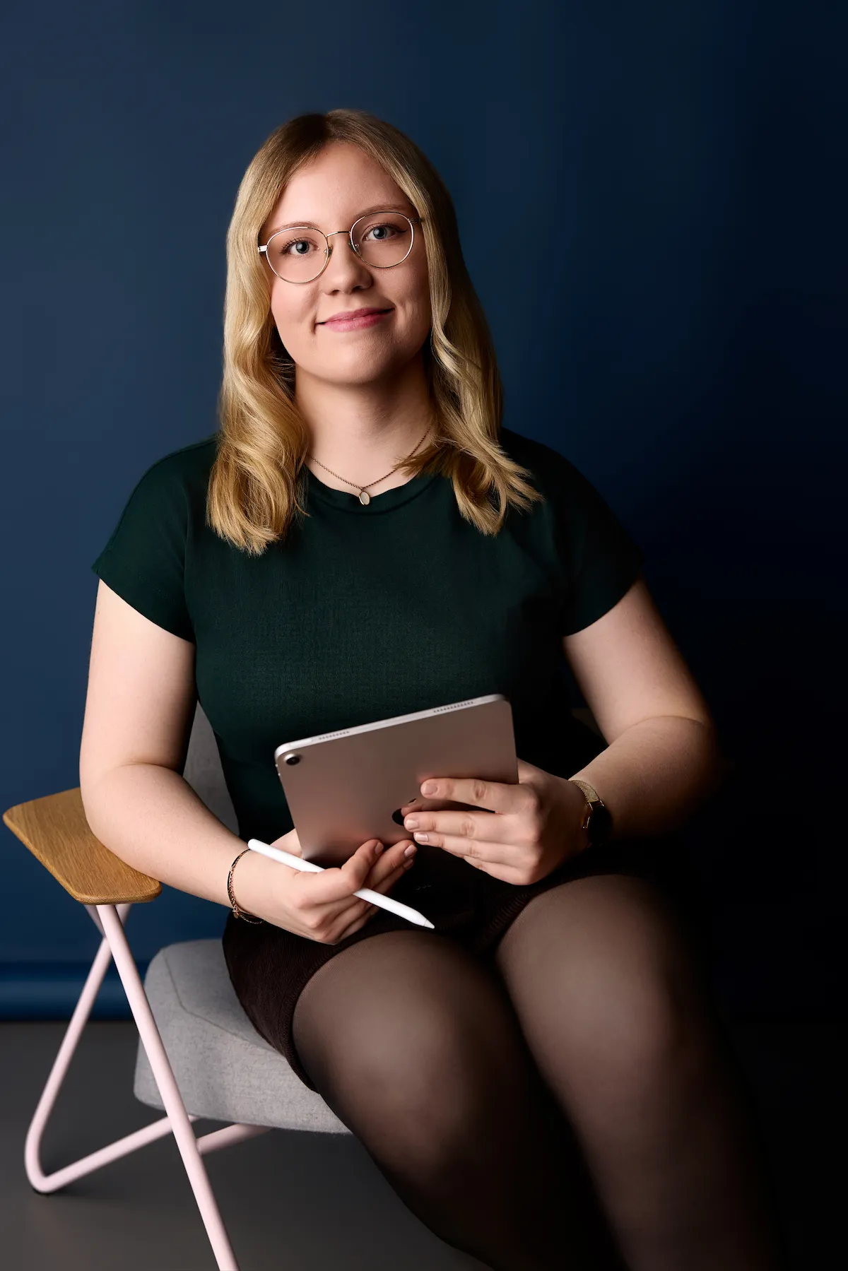 Young woman with glasses sitting on chair holding tablet and stylus against dark blue background.