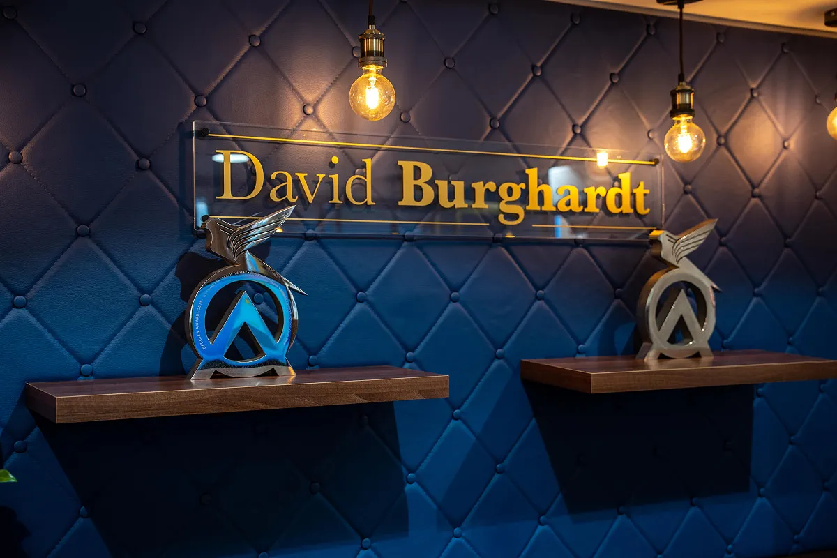 Blue quilted wall with golden David Burghardt sign and two shelves holding winged awards under hanging lights.