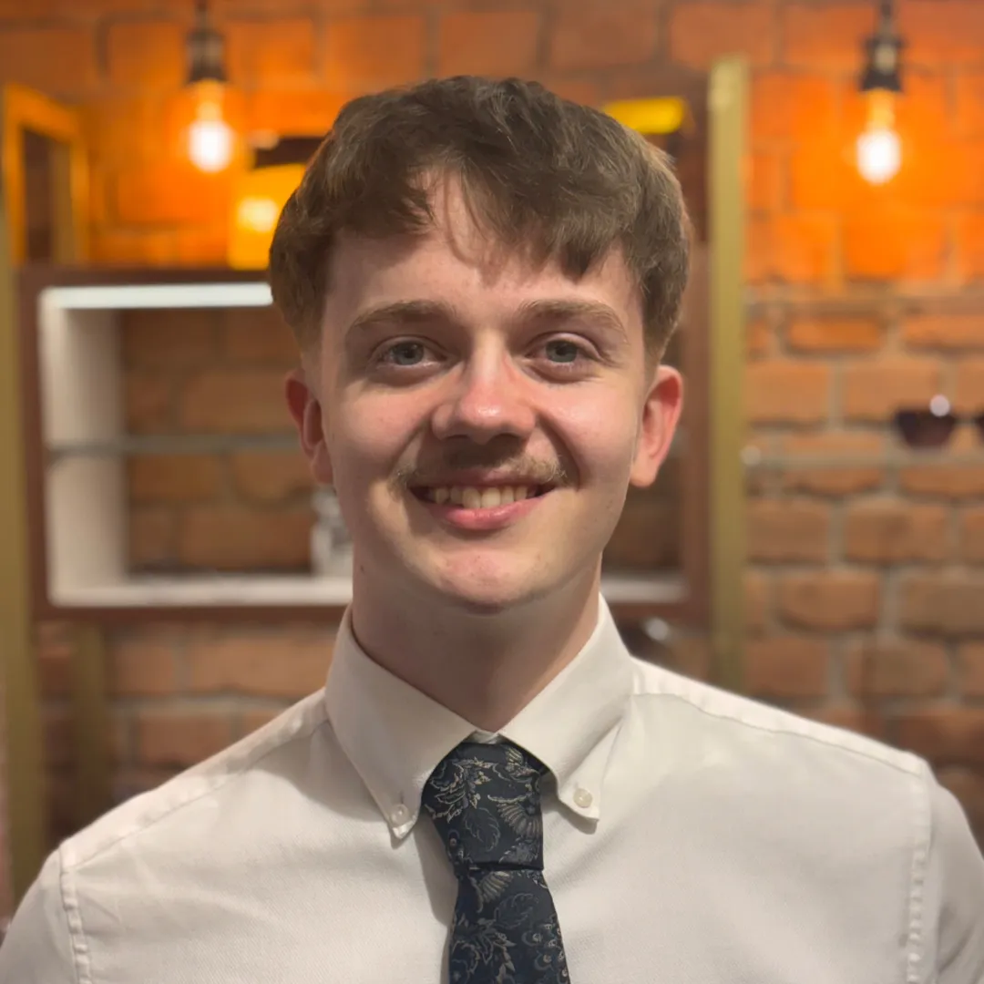 Young man in white shirt and patterned tie smiling in a warmly lit room with brick walls.