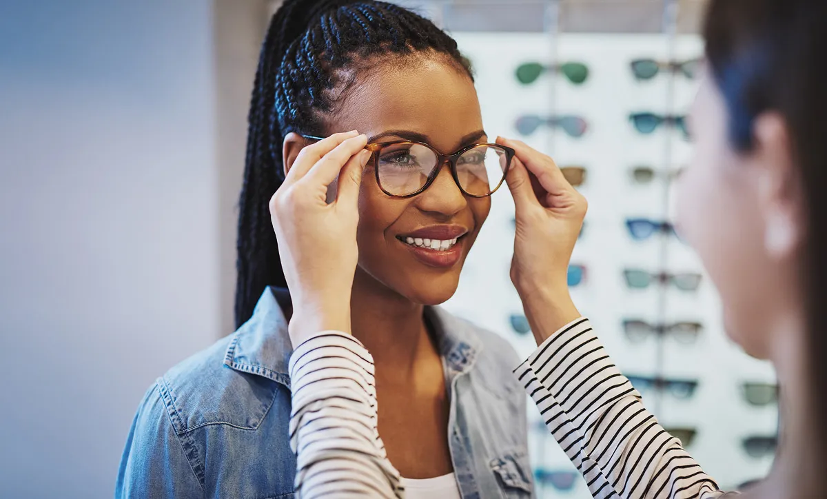 Woman with braided hair smiling as another person helps her try on eyeglasses in an optical store.