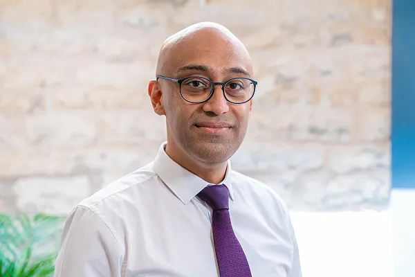 Man with glasses wearing a white shirt and purple tie, smiling slightly against a neutral background.