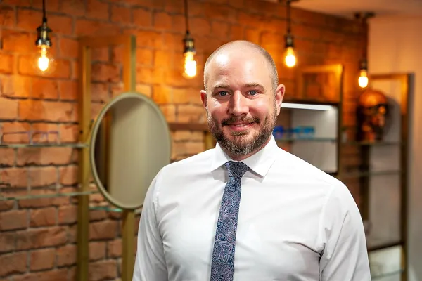 Smiling bald man with a beard wearing a white shirt and patterned tie in a warmly lit room.