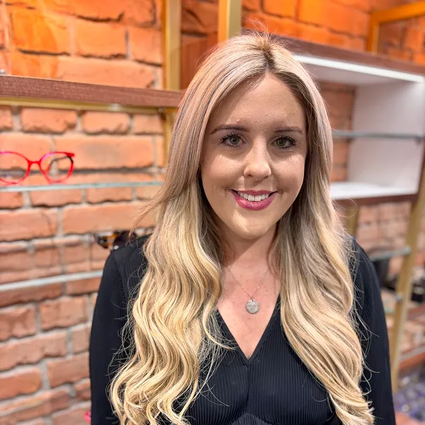 Smiling blonde woman with long wavy hair wearing a black top in a brick-walled room with glasses on shelves.