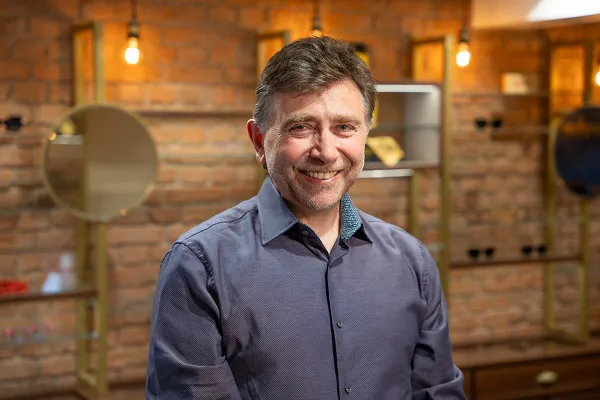 Smiling man with short hair in a blue shirt stands indoors with warm brick wall background.