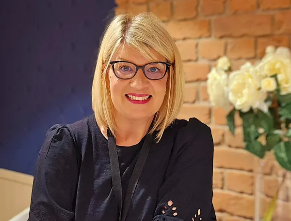 Smiling blonde woman with glasses wearing a black top in front of a brick wall and white flowers.