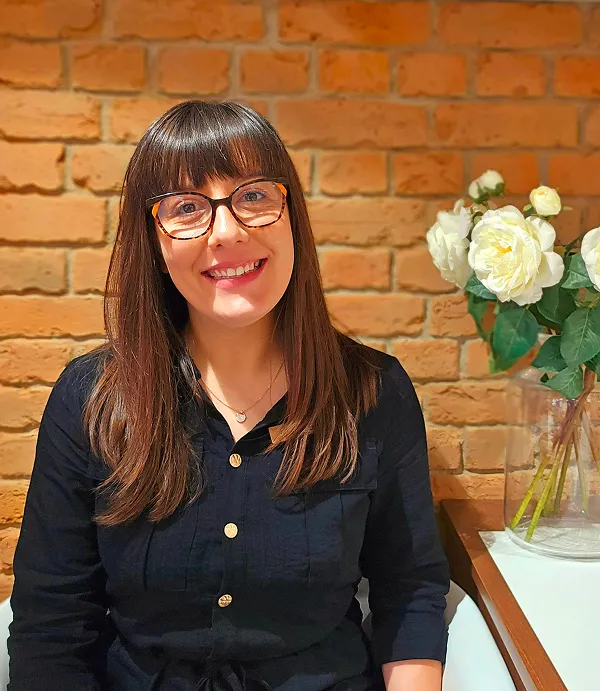 Smiling woman with straight brown hair and glasses sitting against a brick wall with white flowers nearby.