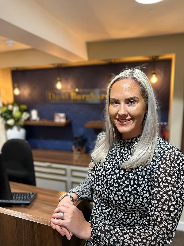 Smiling woman with blonde hair and patterned blouse stands at a wooden reception desk indoors.