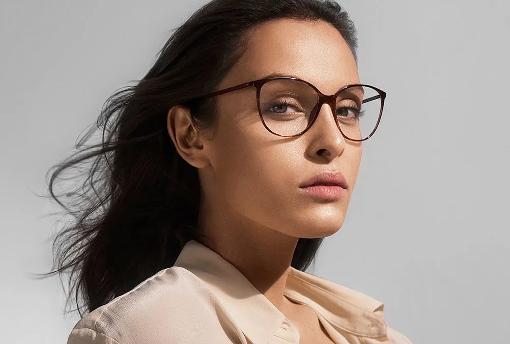 Woman with dark hair wearing stylish brown eyeglasses and a beige top against a gray background.
