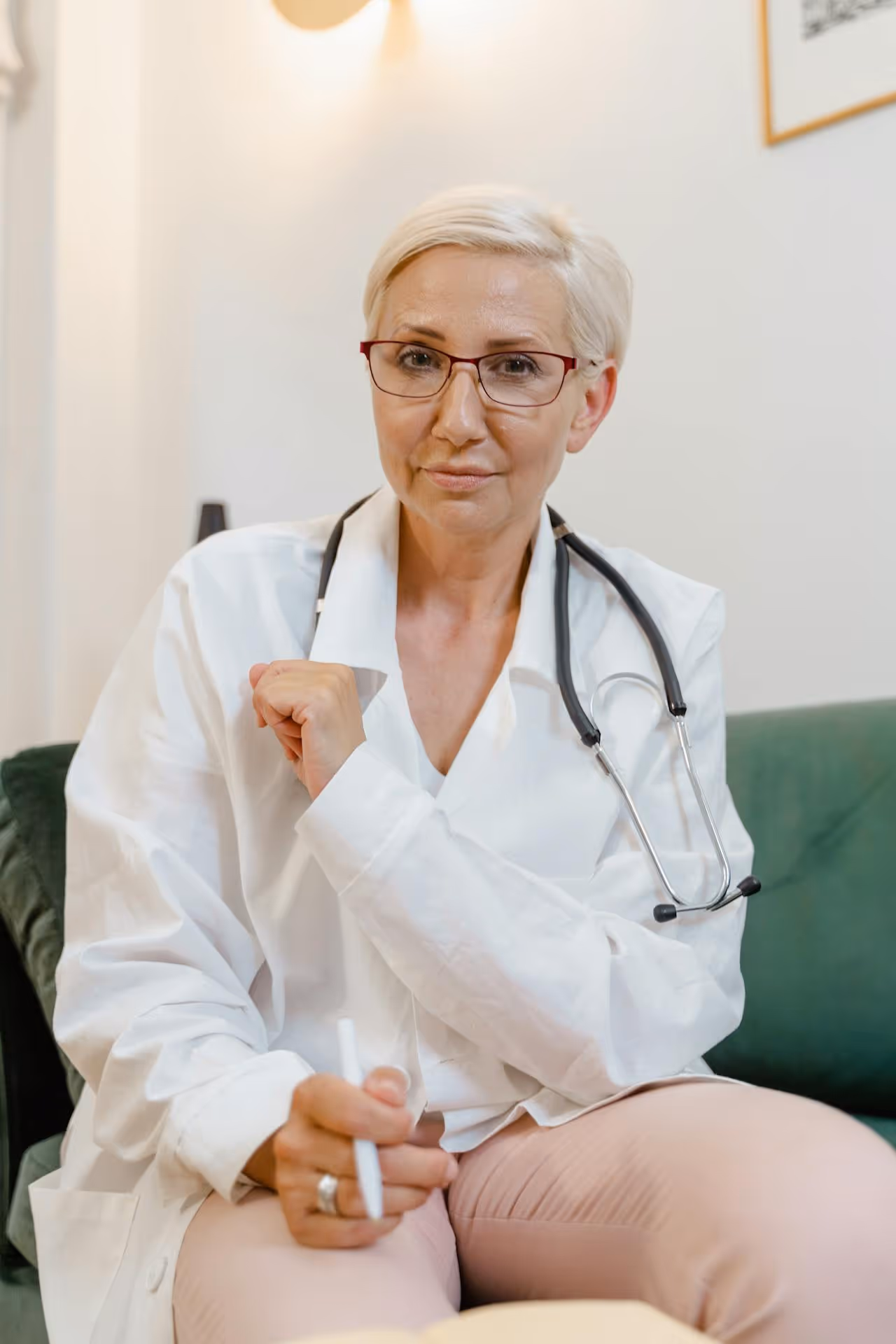 Middle-aged female doctor in white coat with stethoscope sitting on green couch, holding a pen.