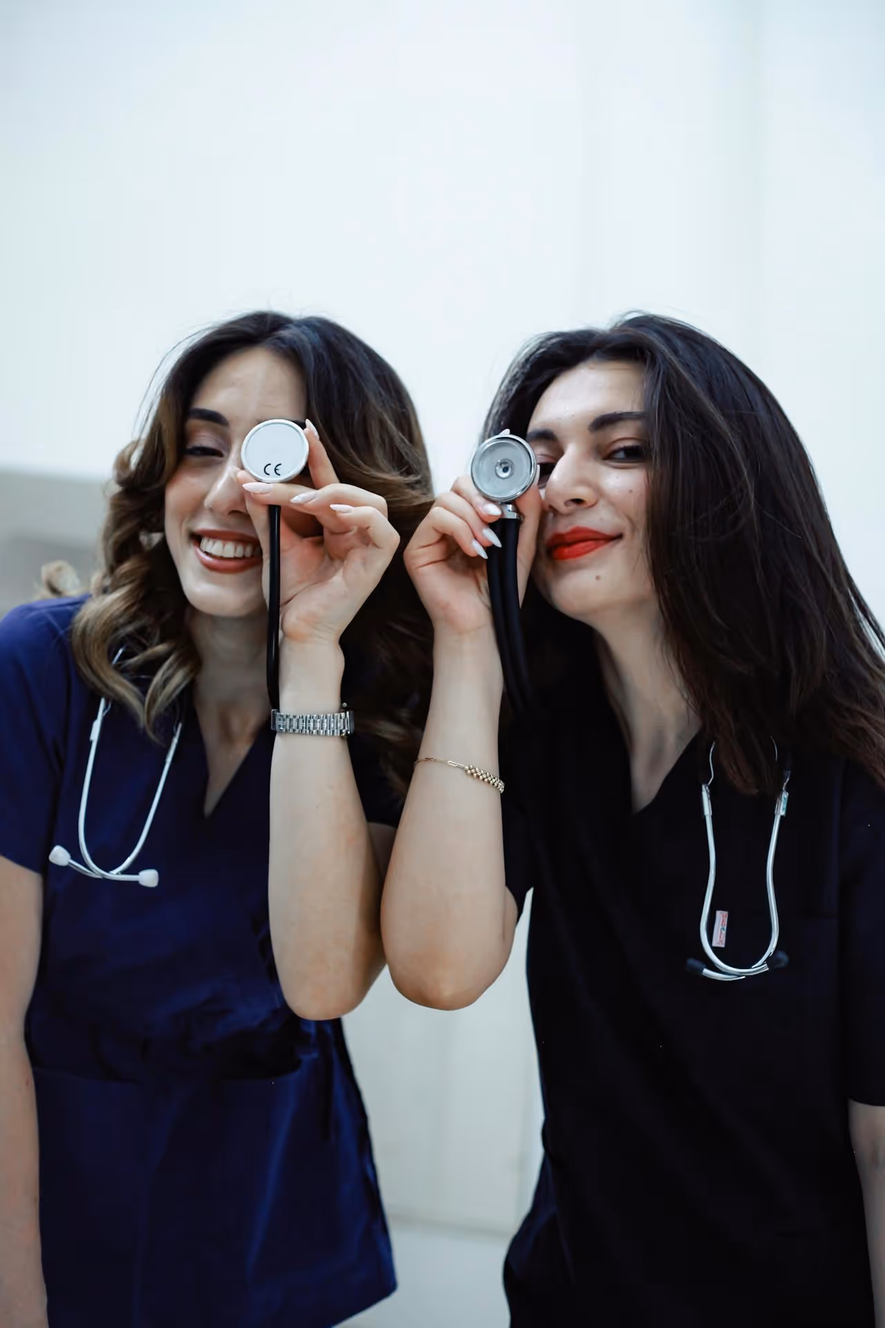 Two female medical professionals smiling and holding stethoscopes up to one eye each like a monocle.