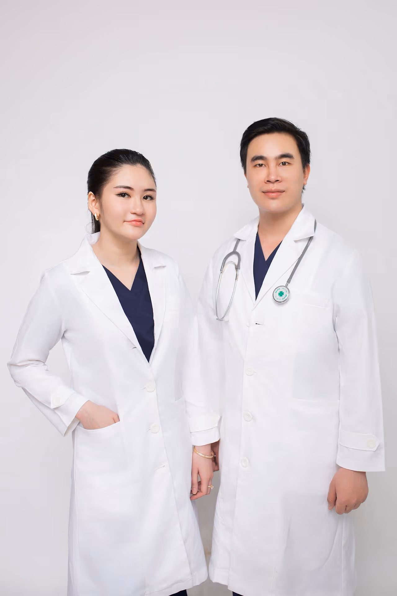Male and female doctors wearing white coats and navy blue scrubs, standing against a plain background, with the male doctor wearing a stethoscope.