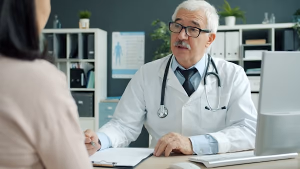 Friendly senior doctor is talking to female patient during appointment in clinic discussing disease and treatment. Communication and medicine concept.