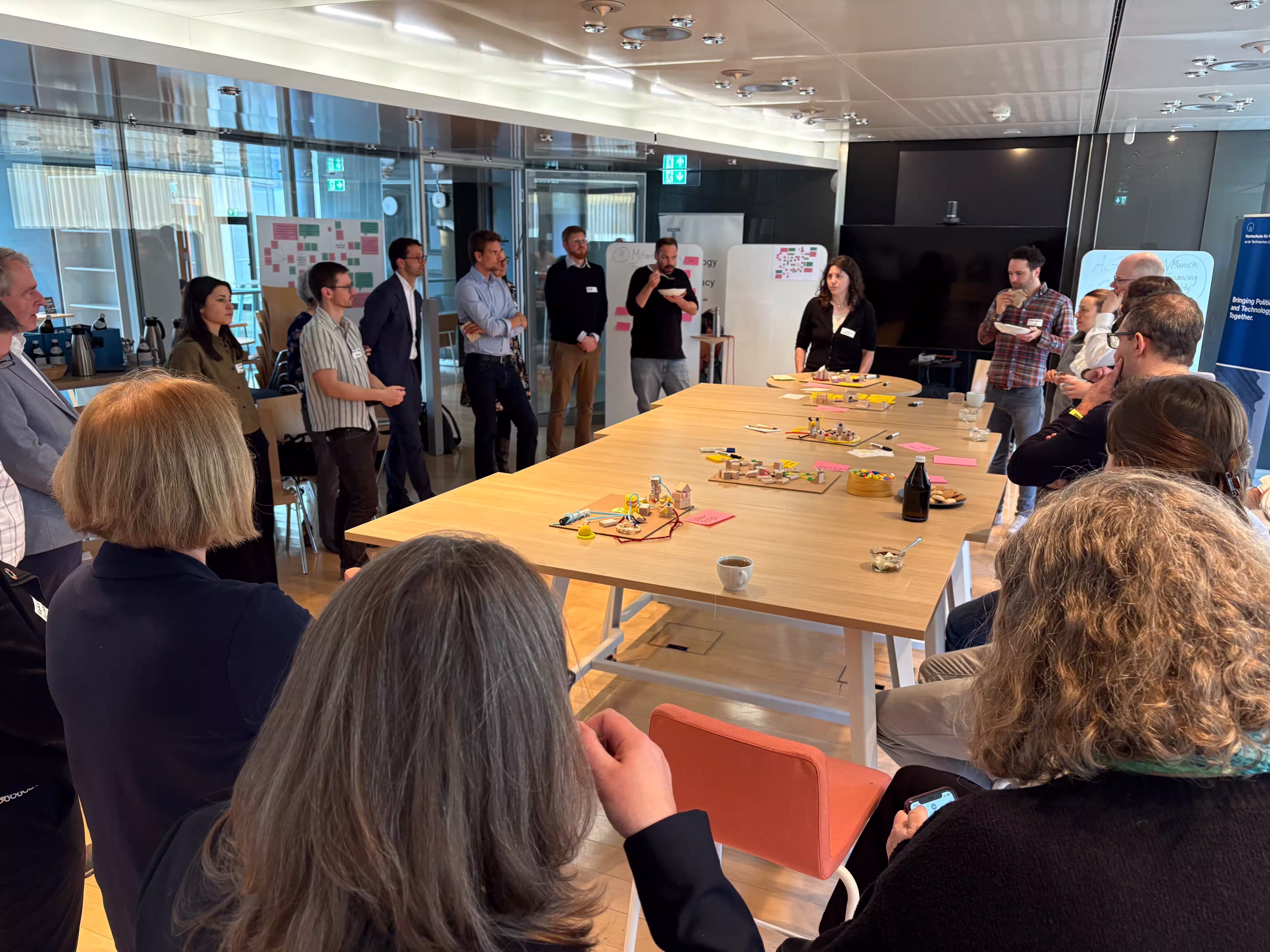 Gruppe von Teilnehmenden steht und sitzt um einen langen Tisch bei einem Workshop der Munich Financing Facility, auf dem Tisch Moderationsmaterial und Notizen.