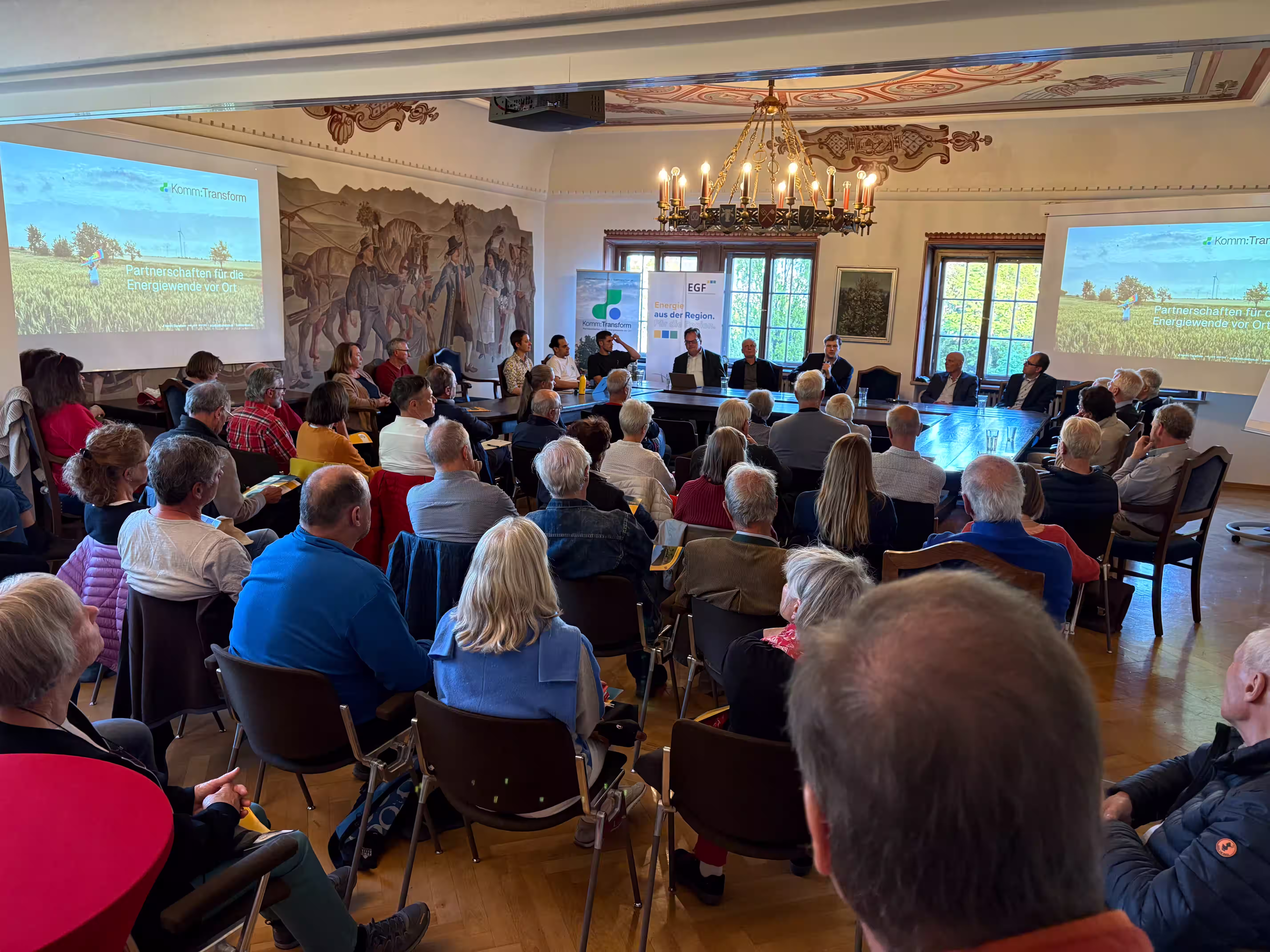 Vollbesetzter Veranstaltungssaal mit historischem Deckengemälde bei einer Komm:Transform-Veranstaltung zum Thema Partnerschaften für die Energiewende vor Ort.