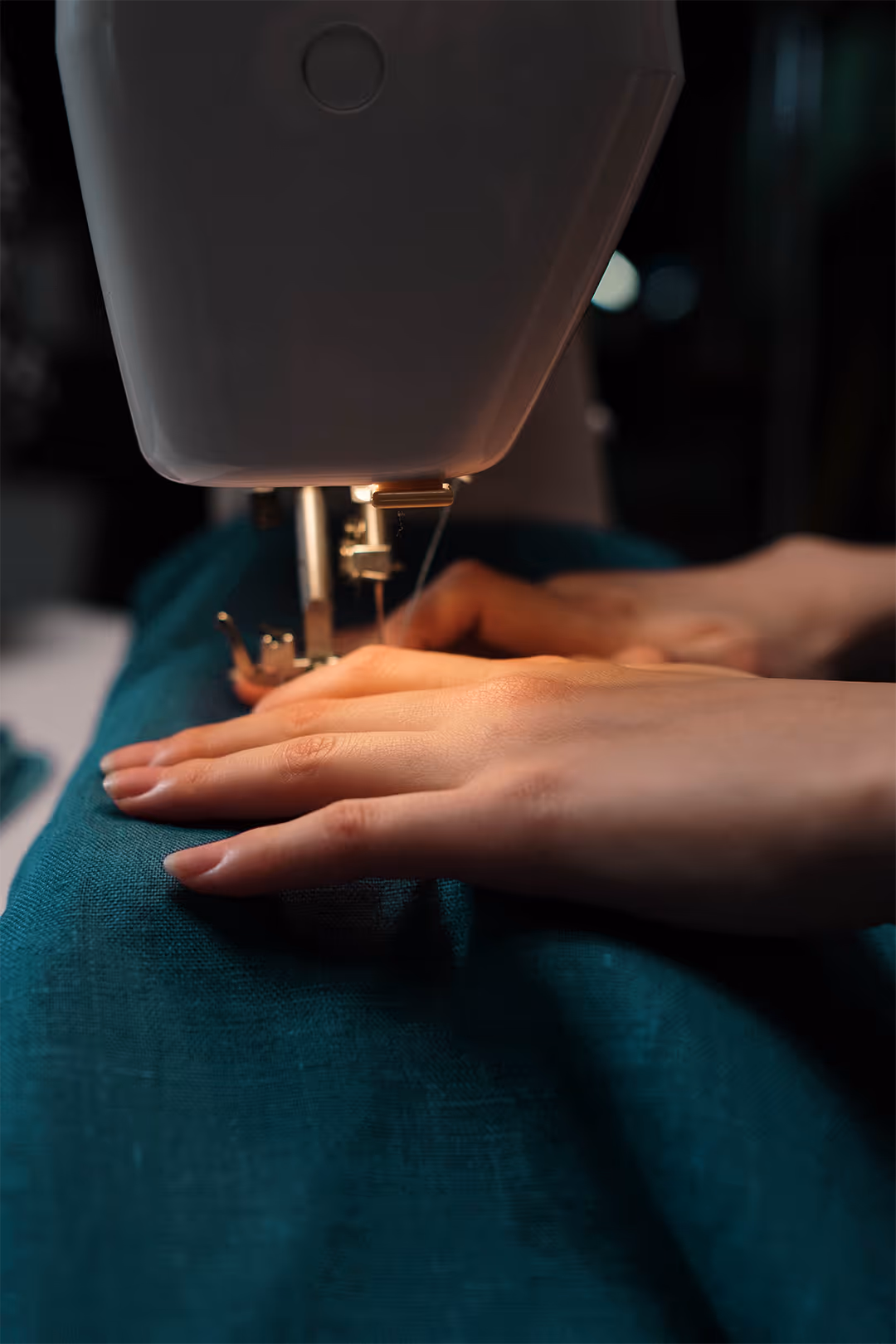 Close-up of hands guiding teal fabric through a sewing machine stitching thread.