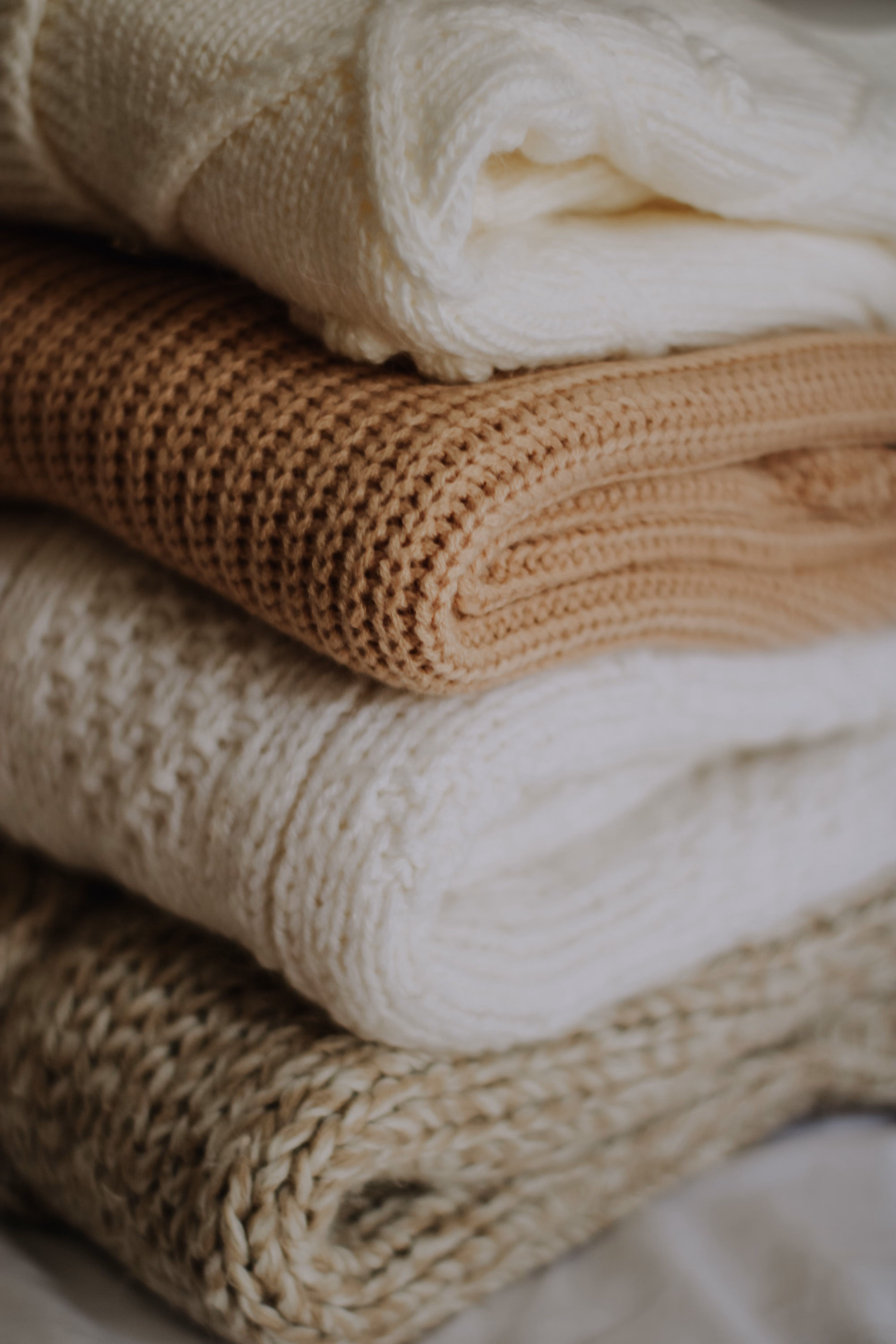 Stack of four folded knit sweaters in beige, white, and brown colors with textured stitches.