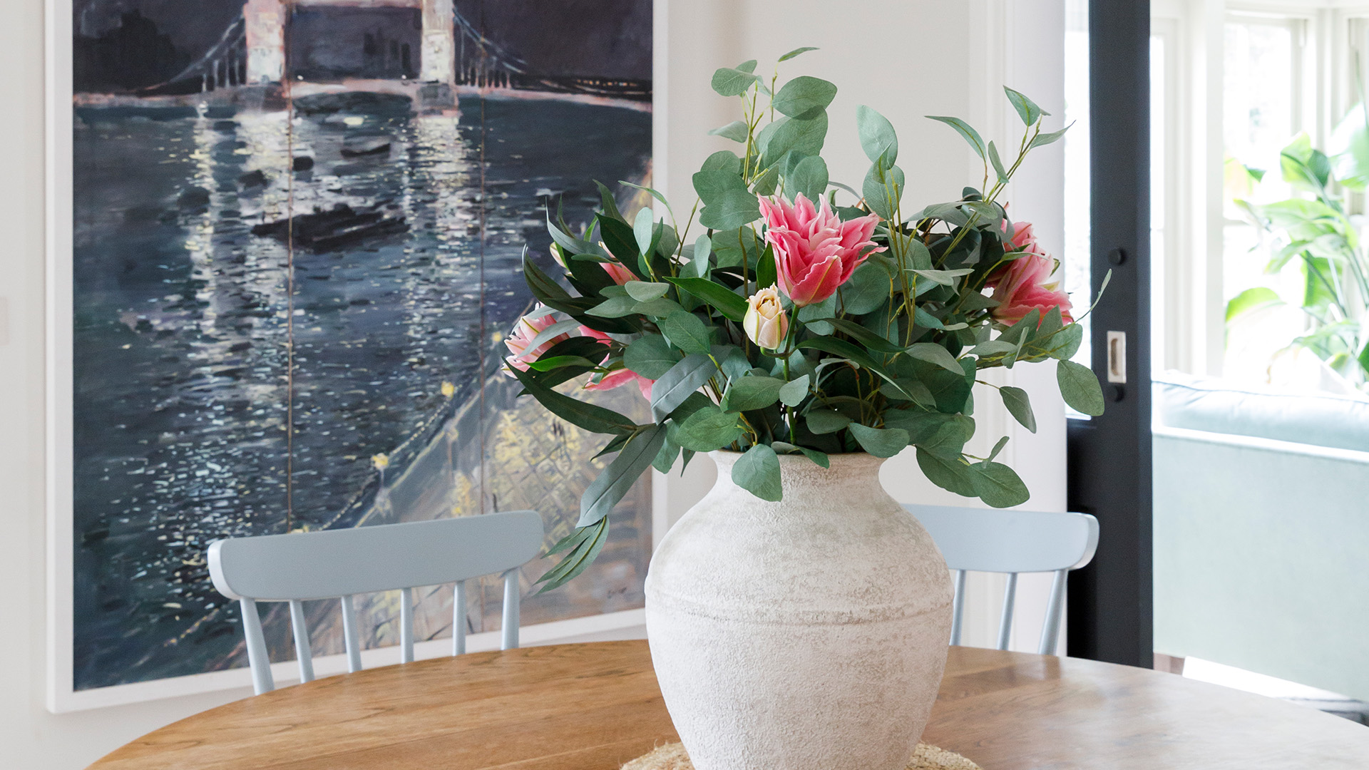Stone vase of fresh flowers on an oak dining table in front of a coastal artwork