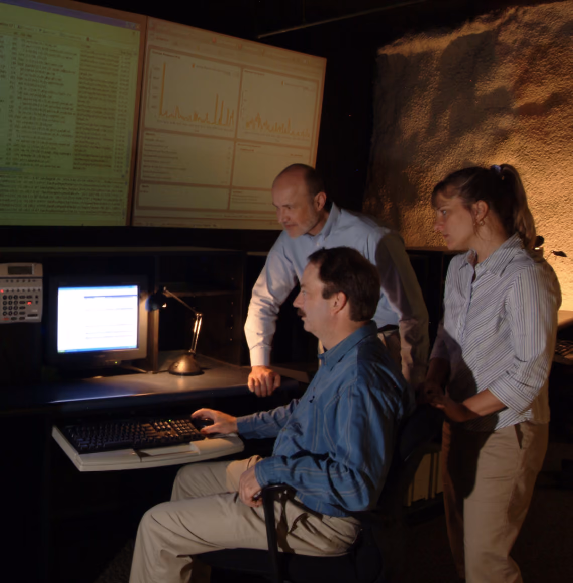 Three people analyzing data on computer screens in a dimly lit room.