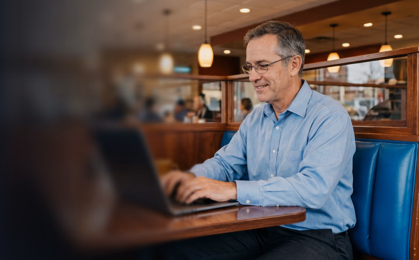 Man in blue shirt and glasses working on a laptop at a wooden table in a café booth.