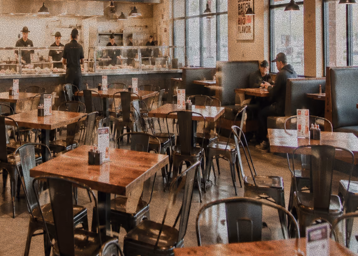Modern casual restaurant with wooden tables, metal chairs, and staff serving behind a glass counter.