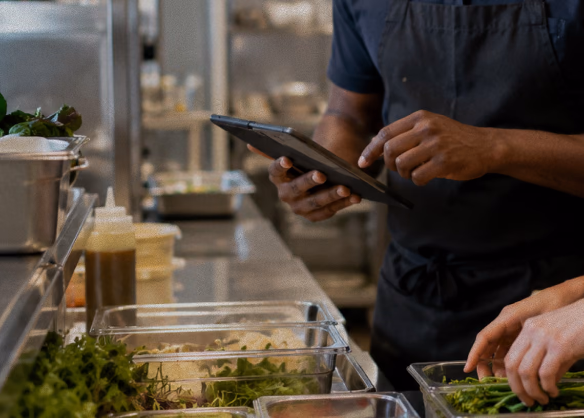 Two people preparing food and using a tablet in a kitchen with containers of leafy greens.