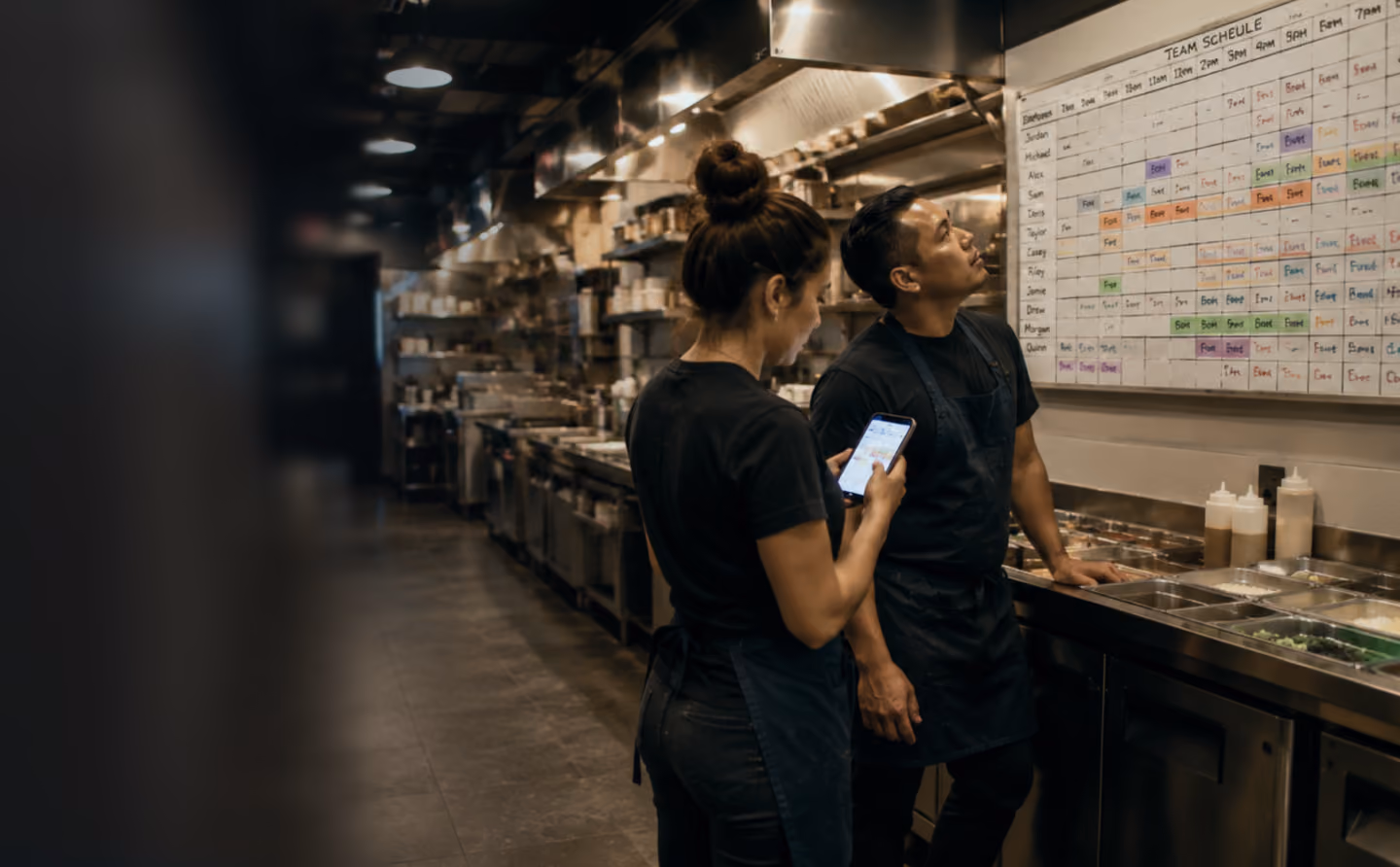 Two kitchen staff in black aprons checking a team schedule on a board in a restaurant kitchen.