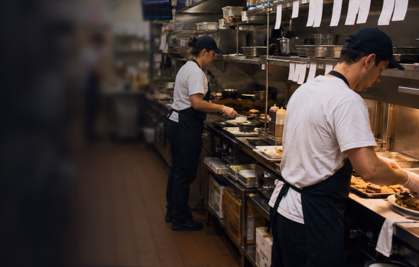 Two chefs wearing white shirts and black aprons working in a busy restaurant kitchen.