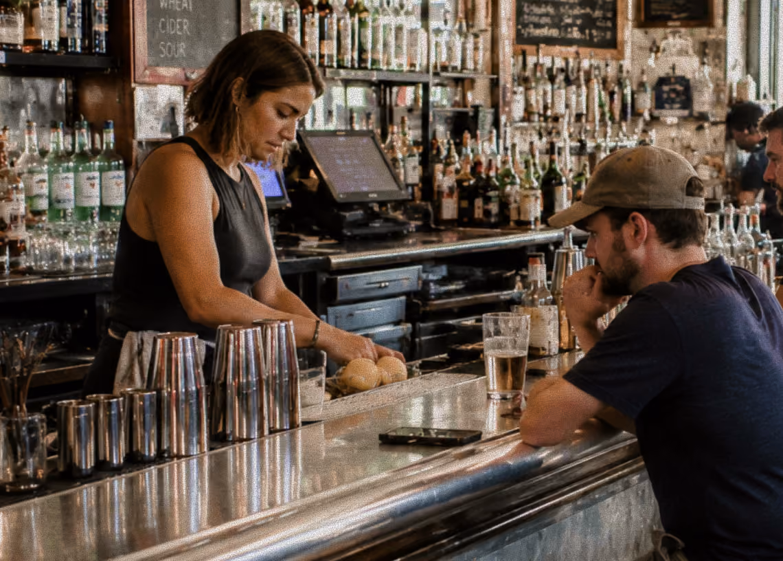 Bartender preparing drinks behind bar as a man in a cap sits with a beer at a busy bar counter.