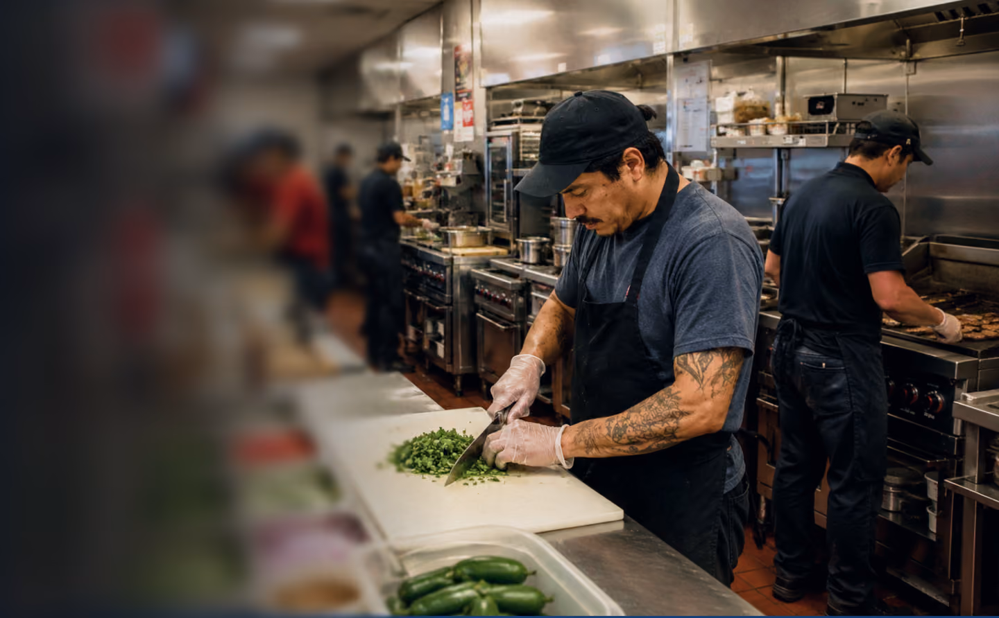Chef wearing black apron and gloves chopping green peppers in a busy commercial kitchen.