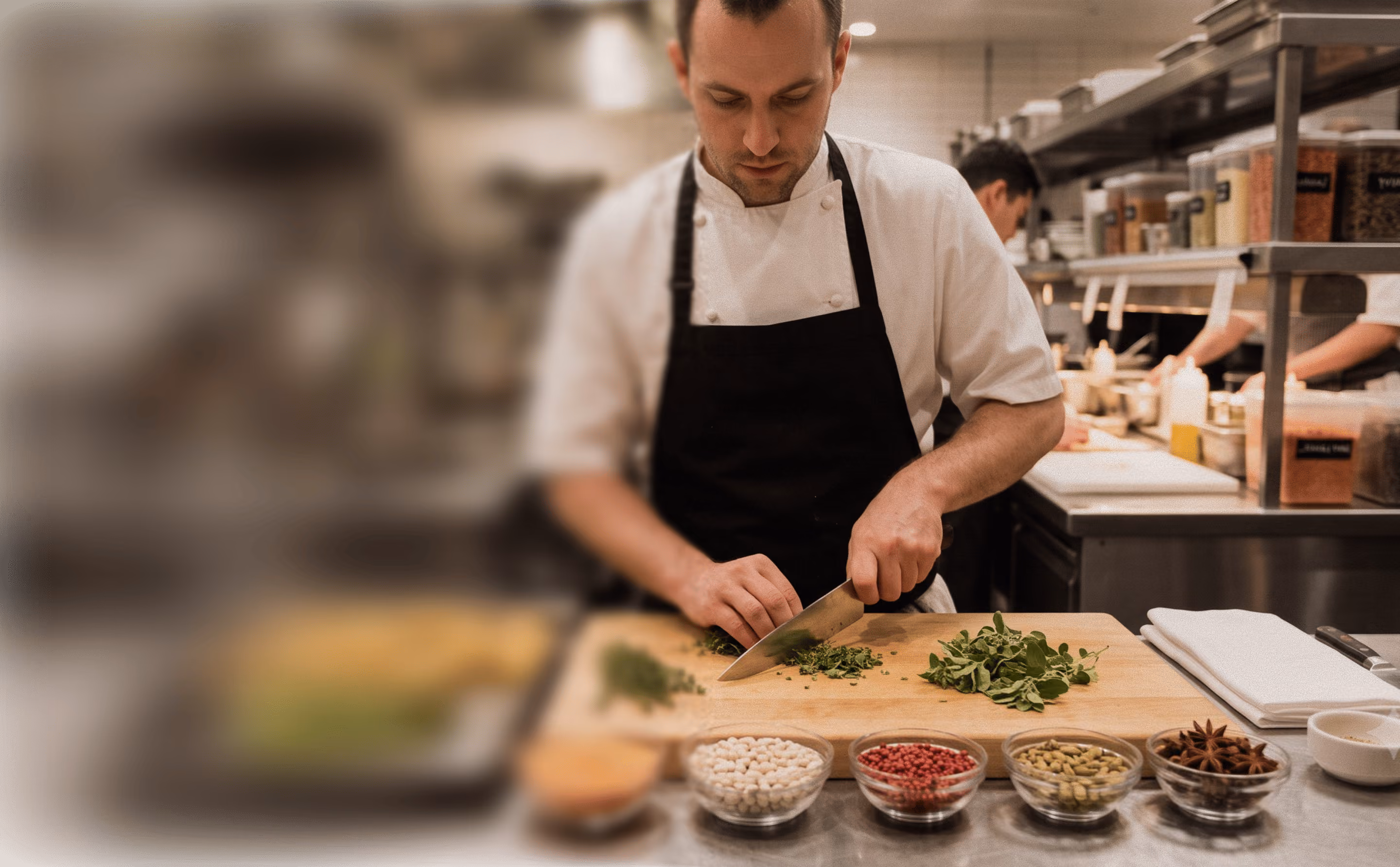 Chef in black apron chopping fresh herbs on a wooden board in a professional kitchen.