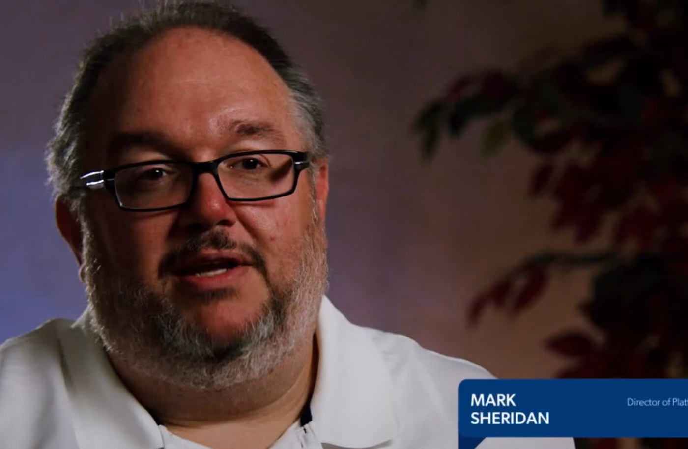 Man with glasses and a beard speaking in a white shirt with a blurred plant background.