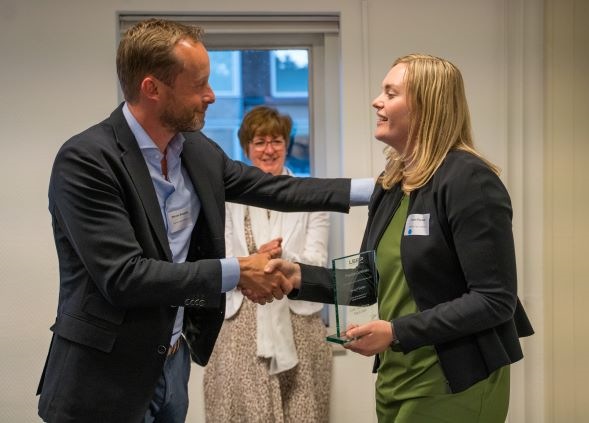 Man schudt vrouw de hand terwijl ze een award ontvangt, met lachende vrouw op de achtergrond.