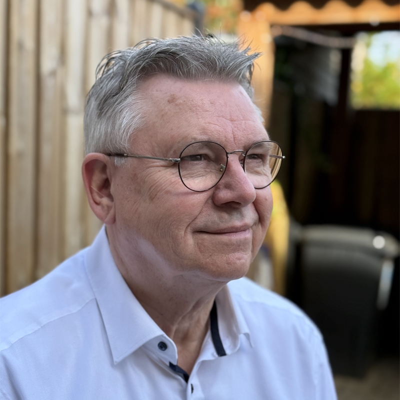 Portret van oudere man met bril en wit overhemd die glimlacht, buiten met houten schutting op de achtergrond.