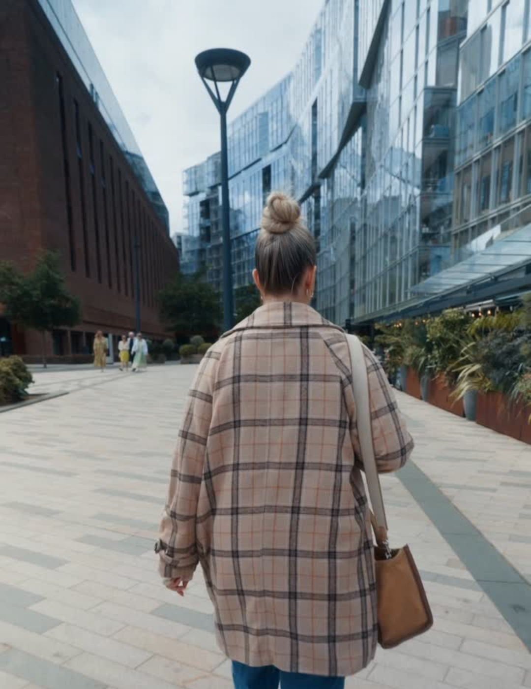 Woman with bun hairstyle wearing a plaid coat walks in an urban area with glass buildings.