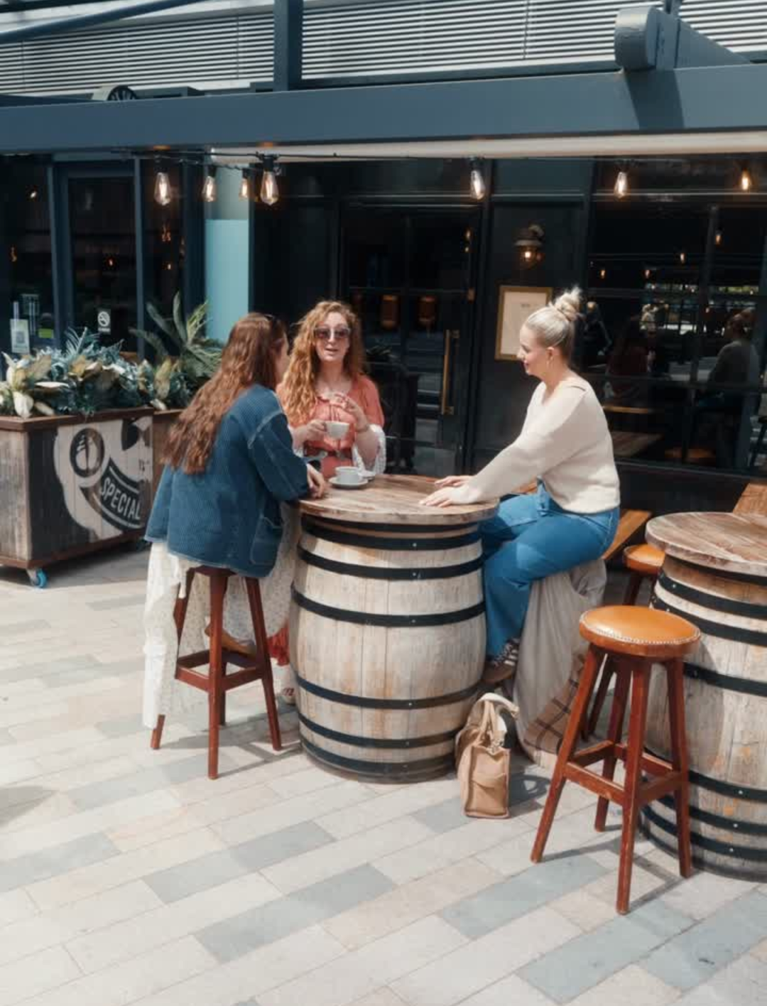 Three women sitting around a wooden barrel table outside a café, chatting and drinking coffee.